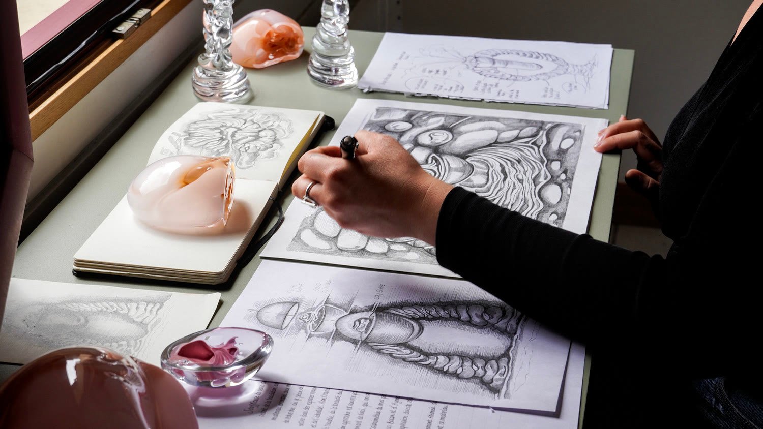 Manon drawing at her desk, glass pieces and a sketchbook around her