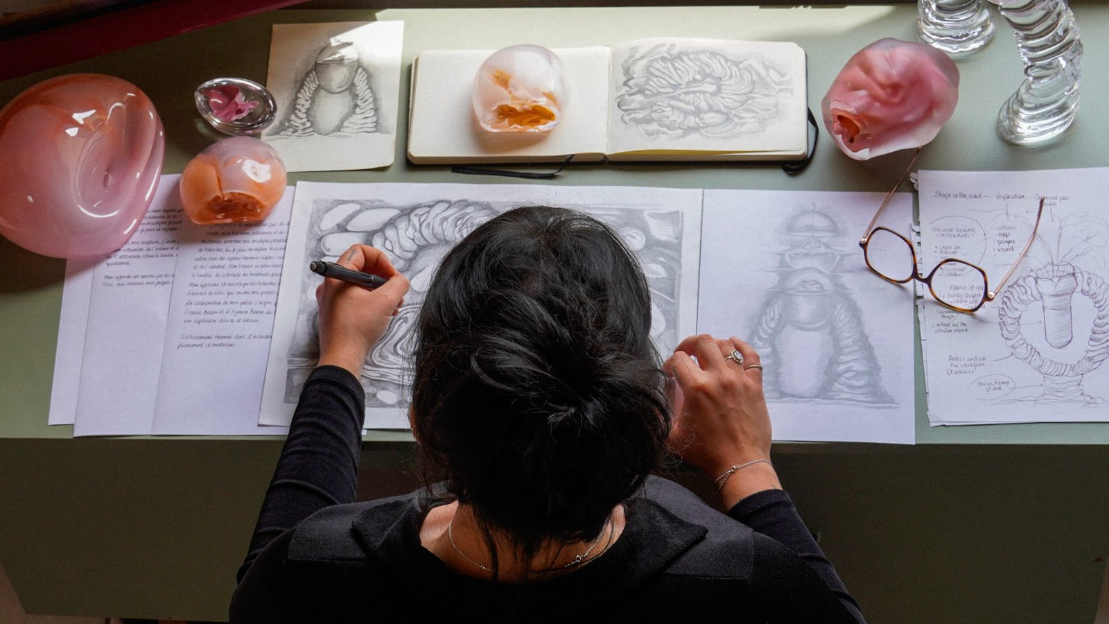 Overhead view of the desk: a drawing in progress, glass pieces and notes