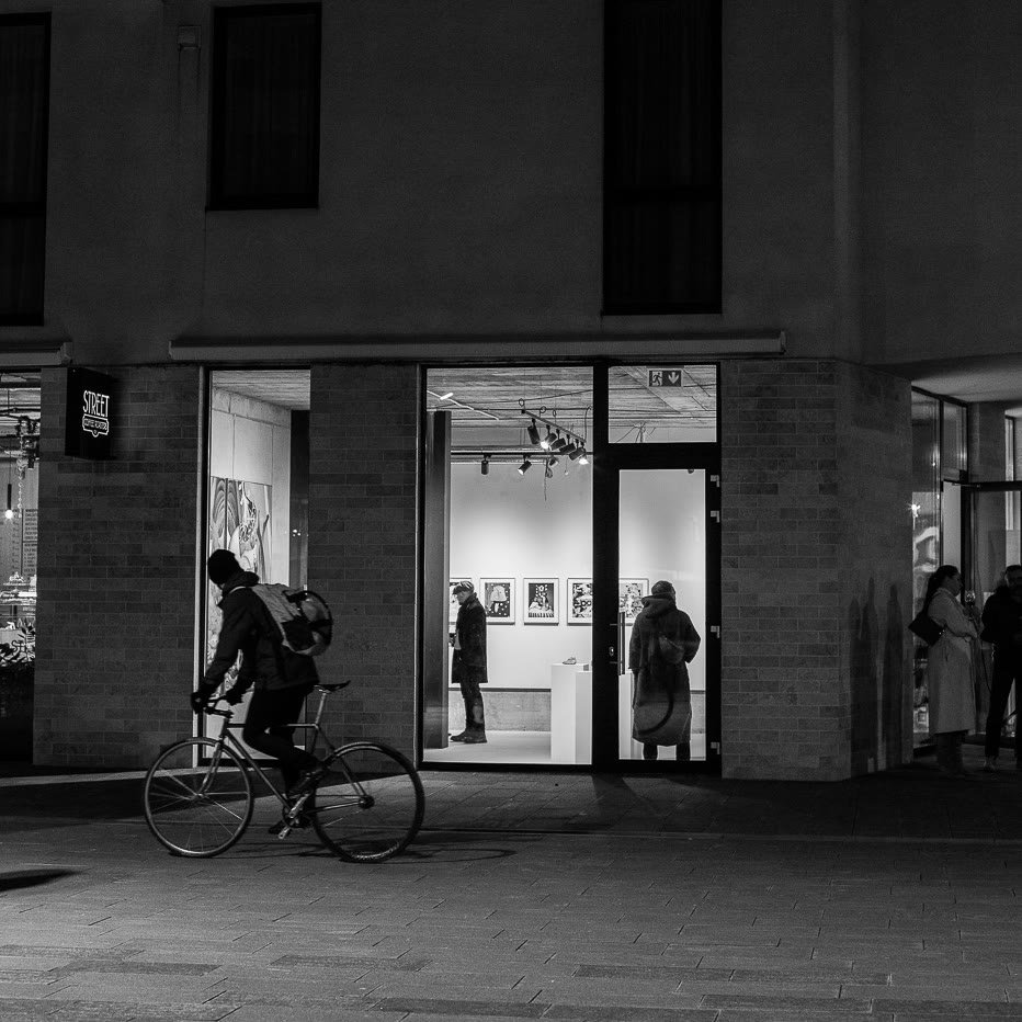The gallery entrance seen from the street, a cyclist passing, in black and white