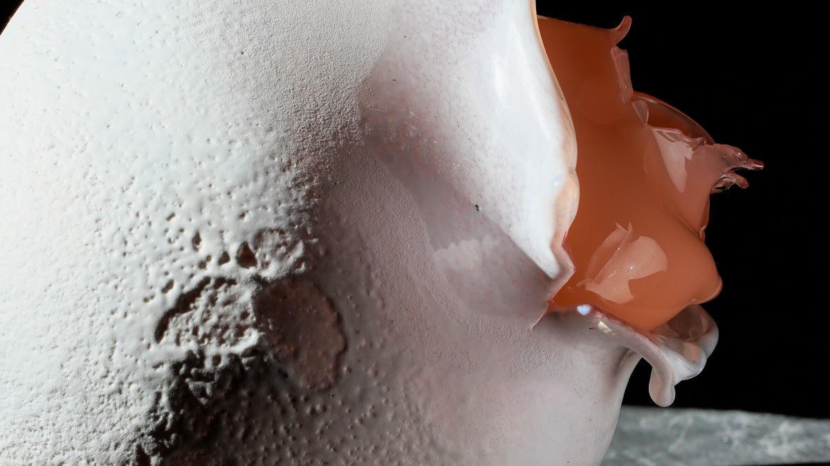A frosted white glass form with a torn opening, glossy coral glass spilling out of it, side-lit on a steel table.