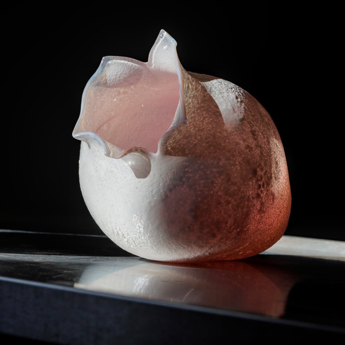 A frosted vessel shading from white to rosy pink, with a jagged translucent pink rim, on a dark reflective plinth.