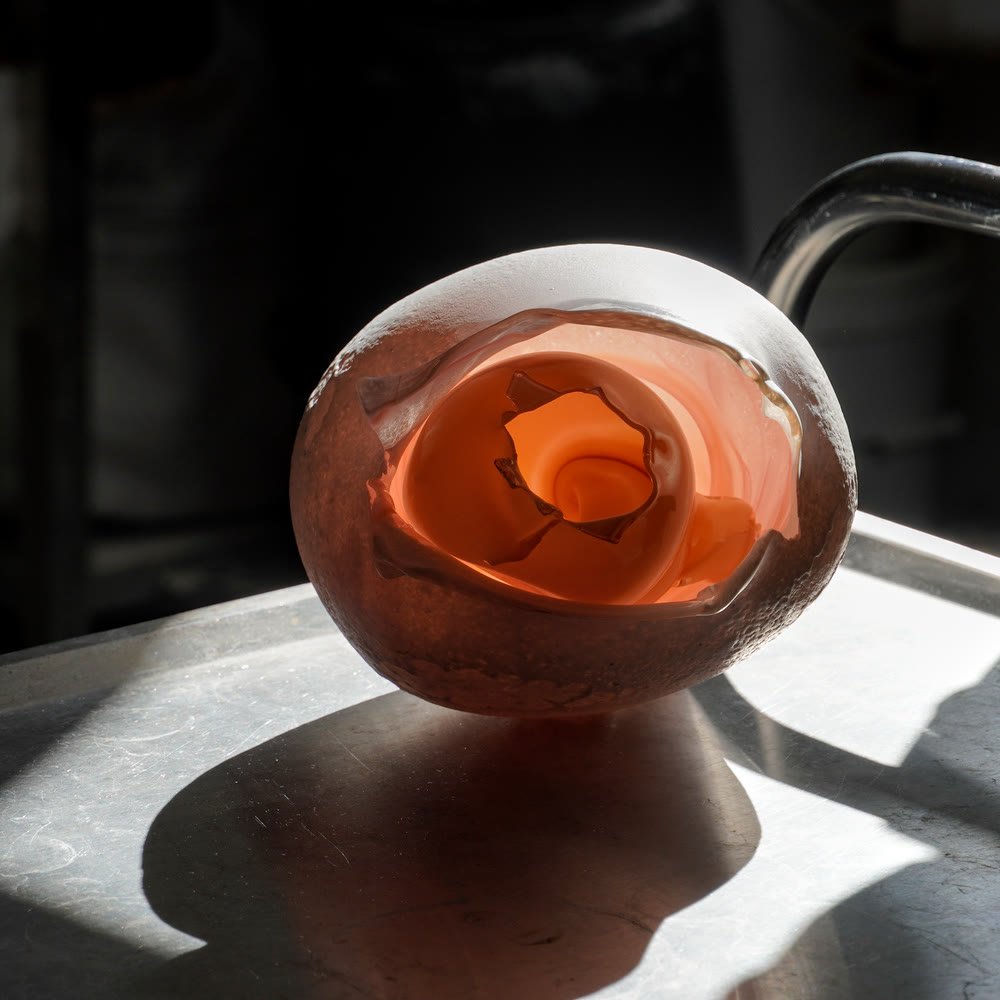 A rounded glass form lit from behind, its spiralling orange core glowing through the frosted skin and casting a warm shadow.